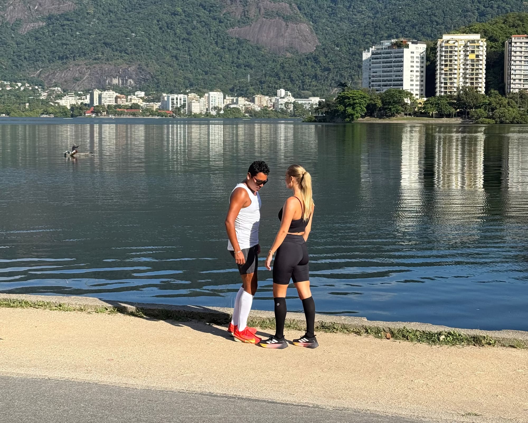 Man och kvinna står vid en sjöpromenad iförda The Pairs kompressionsstrumpor, berg i bakgrunden