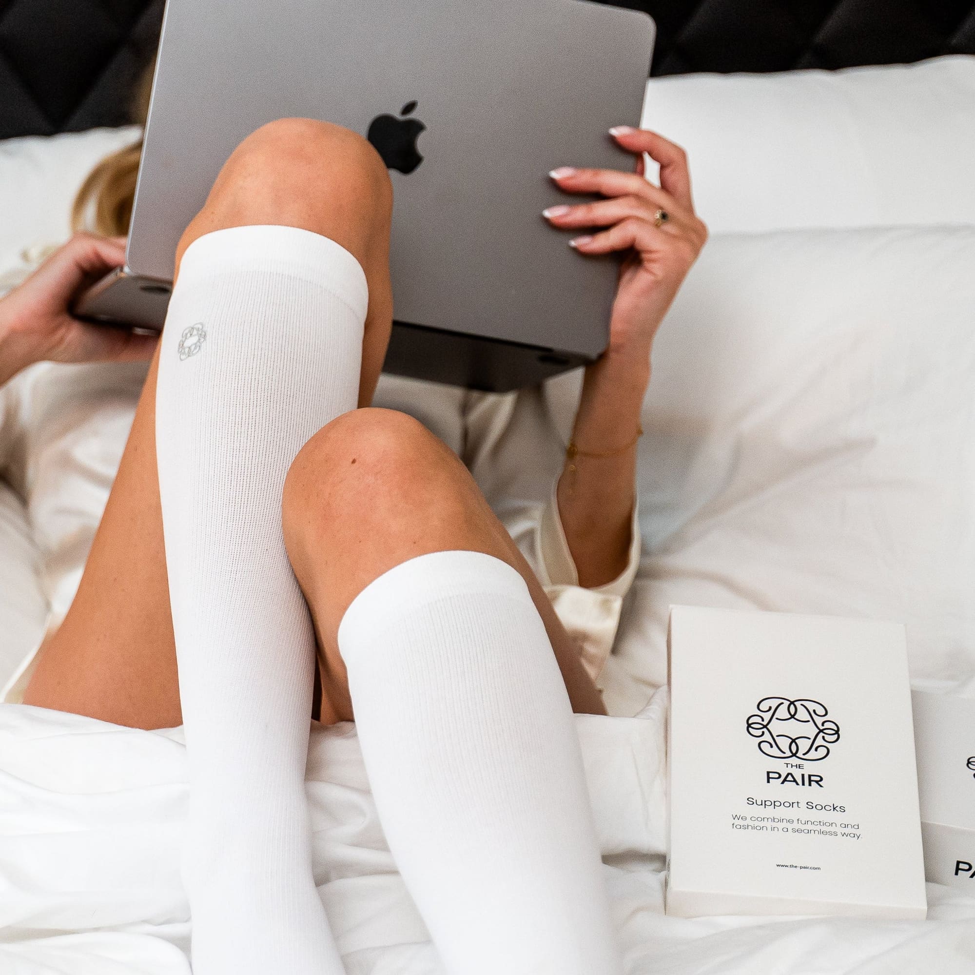 Kvinna i sängen med vita knästrumpor och laptop, The Pairs produktförpackning bredvid